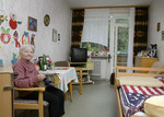 Eine Bewohnerin sitzt auf einem Stuhl in ihrem Zimmer. Neben Stühlen befinden sich in ihrem Zimmer, ein Tisch, ein Bett, ein Fernseher und Schränke.